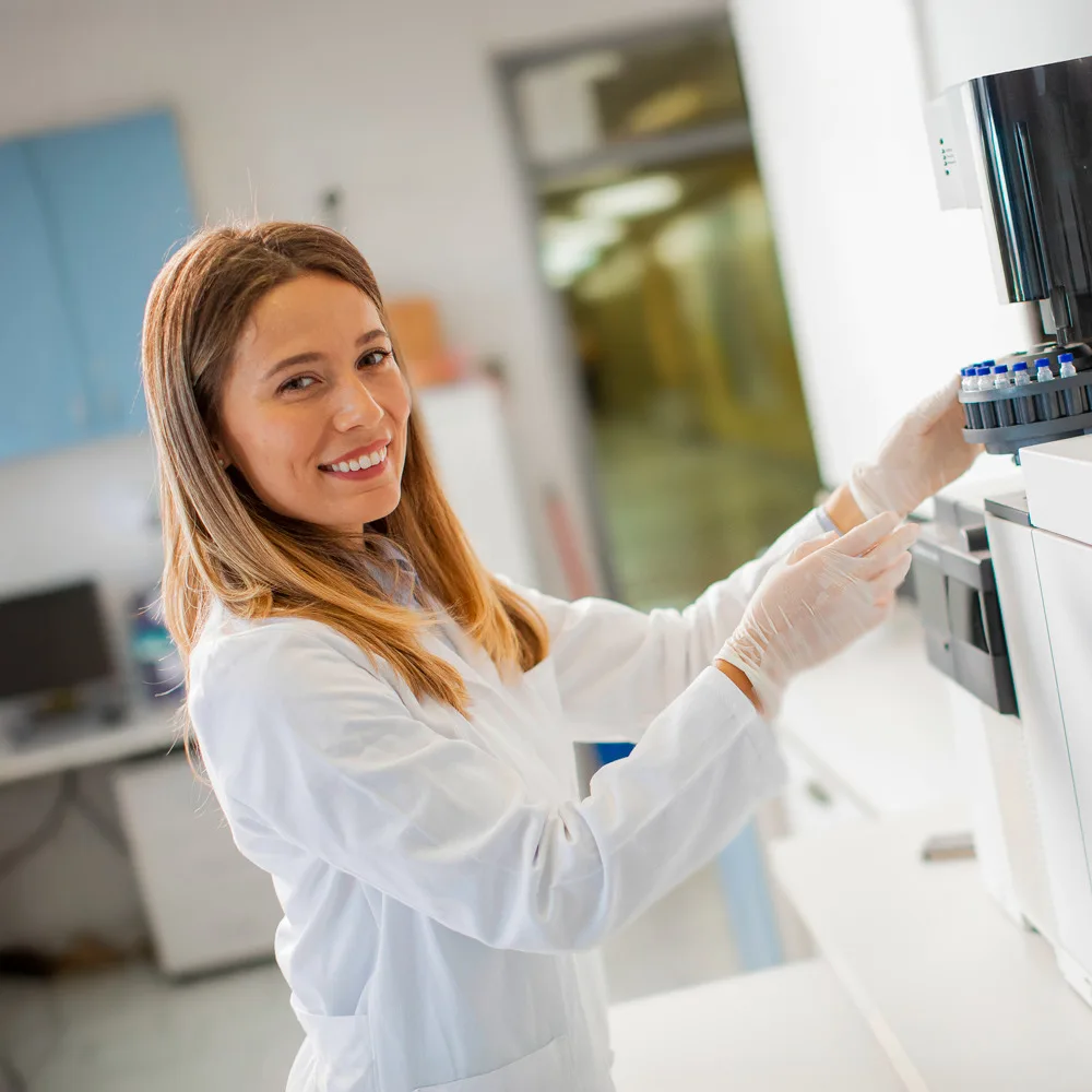 Imagem mostra uma mulher branca sorrindo, ela usa um jaleco branco enquanto manuseia uma maquina