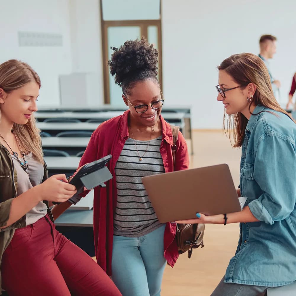 Imagem mostra 2 mulheres olhando para a tela do notebook sorrindo e uma para a tela de um tablet