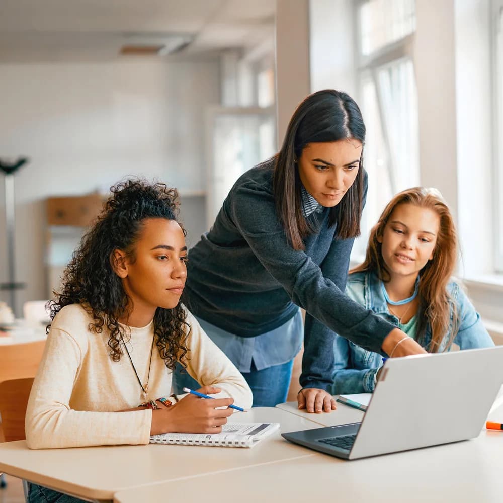 Imagem mostra 3 mulheres olhando para a tela do notebook, 2 estão sentadas e a do meio está em pé apontado para a tela