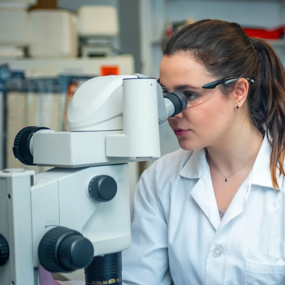 Imagem mostra uma mulher branca de jaleco branco usando um óculos para manusear um microscópio
