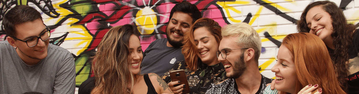 Diversas pessoas olhando para um celular e sorrindo, logo ao fundo há um muro colorido com grafite