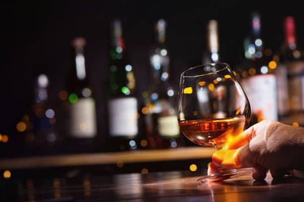 Uma mão segura um copo de conhaque ou uísque em um bar, com garrafas de bebida desfocadas ao fundo..jpg Uma mão segura um copo de conhaque ou uísque em um bar, com garrafas de bebida desfocadas ao fundo..jpg