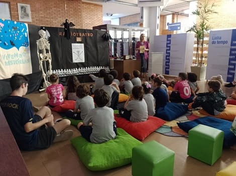 Foto de diversas crianças sentadas ao chão enquanto prestam atenção na história sendo contada por uma professora.